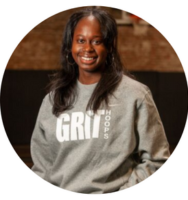 New York Grit Coach Cherkira Lashley, posing in their gray and white sweatshirt, one hand in pocket and smiling confidently against a basketball court backdrop for their official coach photo.